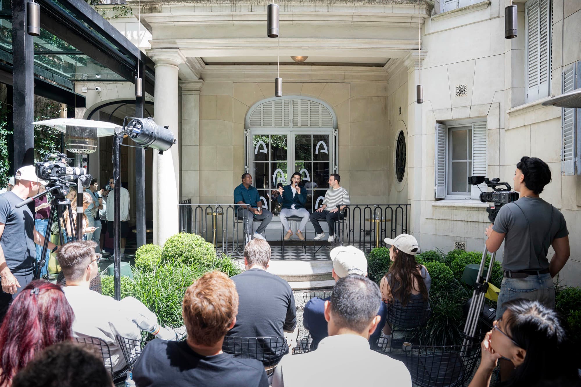 A panel on a terrace stage, filmed by camera crew, with an audience seated in front