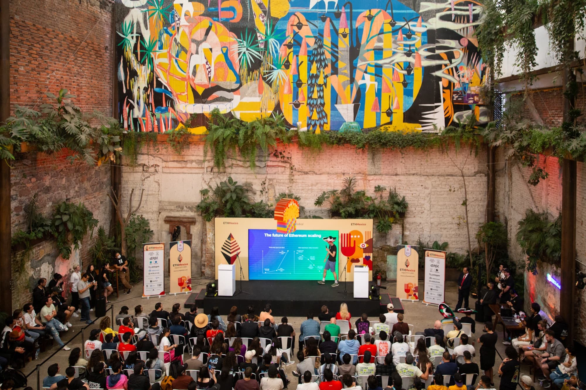 A crowd facing a stage beneath a large mural at ETH Mexico