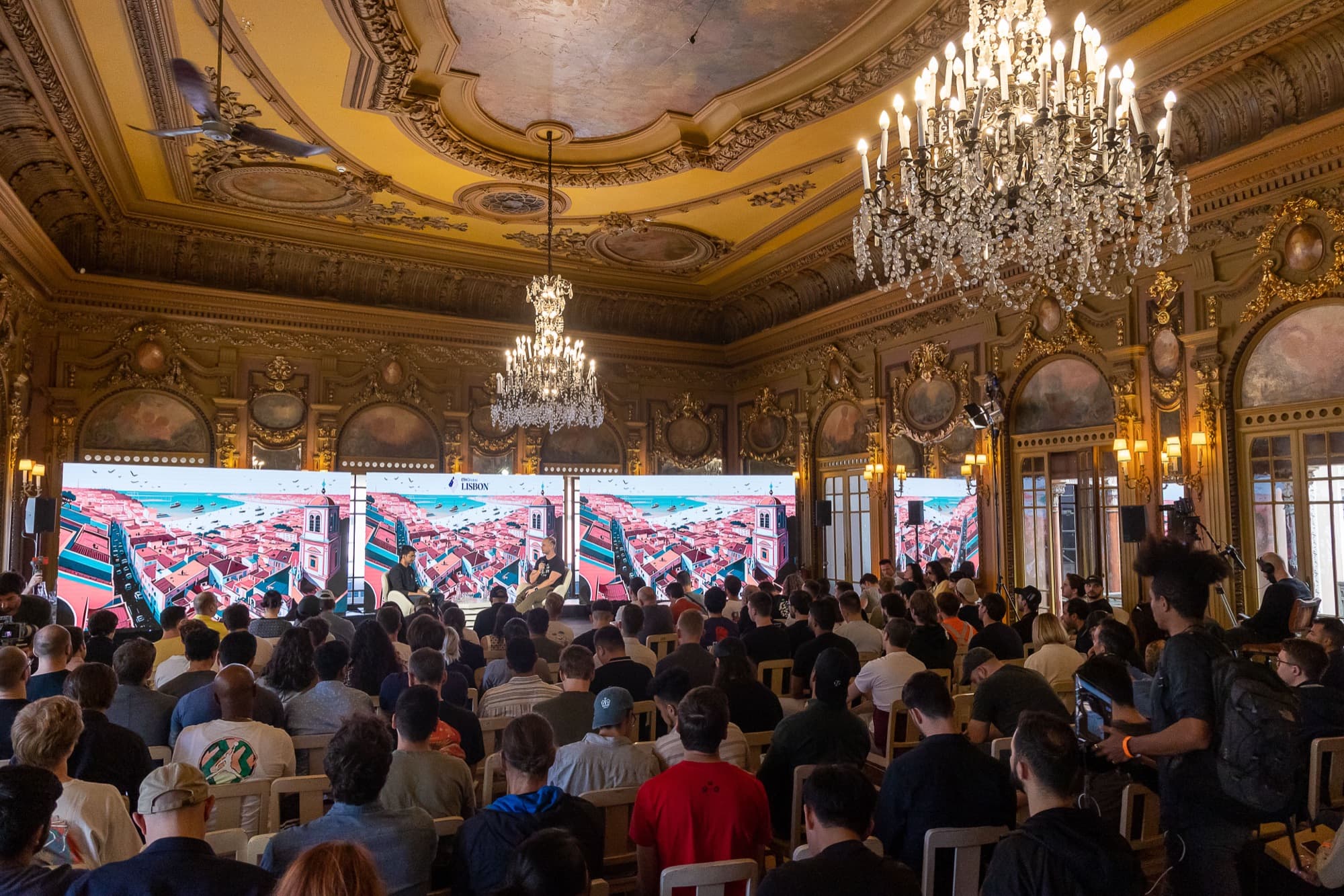 A packed ballroom in Lisbon under chandeliers, screens across the stage