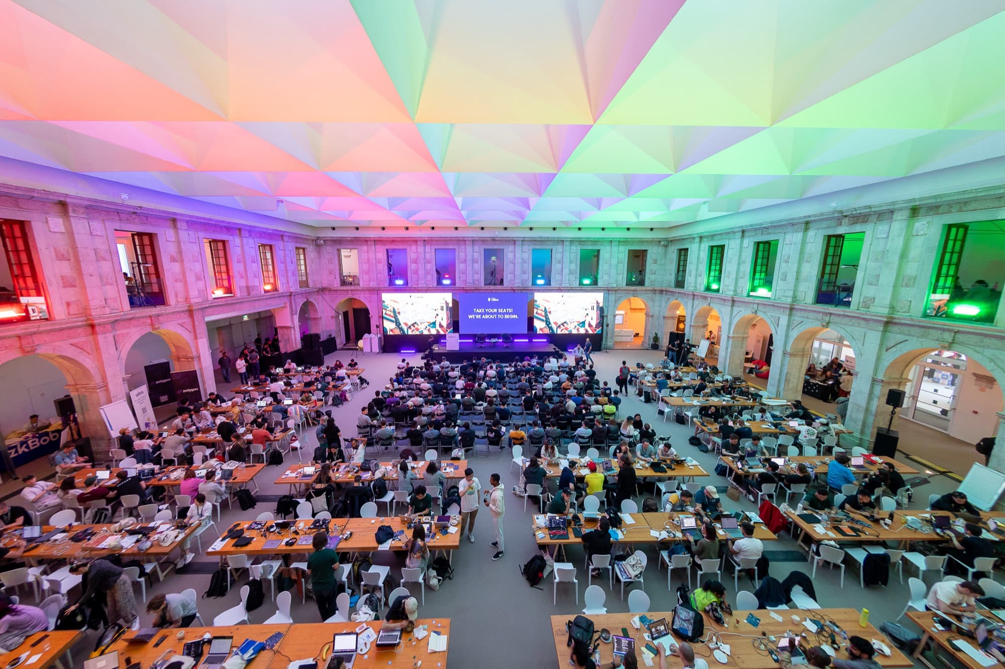 A hackathon hall in Lisbon under a rainbow-lit ceiling, rows of attendees at work